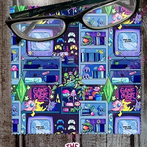 May include: A colorful microfiber cleaning cloth with a retro video game theme. The cloth features a repeating pattern of pixelated characters, game consoles, and controllers. The text "GAME OVER" appears in neon pink and blue.