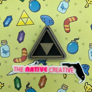 May include: A collection of colorful, cartoon-style stickers and pins on a light green background. The designs include a Triforce symbol, bombs, bottles, and a boomerang. The text "THE NATIVE CREATIVE" is also visible.