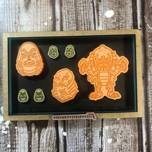 May include: A set of five wooden creature from the black lagoon themed pins. The set includes a large pin of the creature's head, a smaller pin of the creature's head, and three smaller pins of the creature's face. The pins are orange and green and are displayed in a wooden frame. The frame has a teal blue mat and a gold border. The text "@thecreativecreativefl" is printed on the bottom of the frame.
