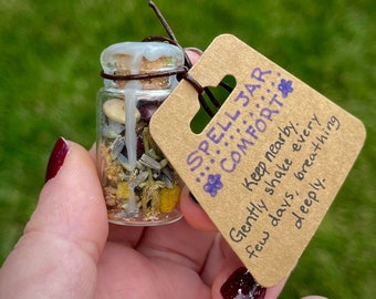May include: A small glass jar with a cork stopper and a brown paper tag. The jar is filled with dried herbs and flowers. The tag reads "SPELL JAR: COMFORT. Keep nearby. Gently shake every few days, breathing deeply."