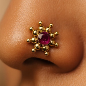 Puede incluir: Un piercing de nariz dorado con una gema central redonda de color rosa intenso. El piercing presenta un diseño de rayos de sol con pequeños detalles esféricos. La joya se encuentra sobre la piel de una persona.
