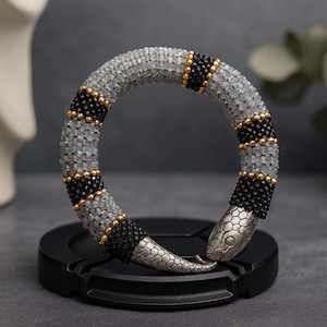 Può includere: Un braccialetto a forma di serpente fatto di perline grigie, nere e dorate. La testa e la coda del serpente sono in metallo argentato. Il braccialetto è esposto su una superficie nera.