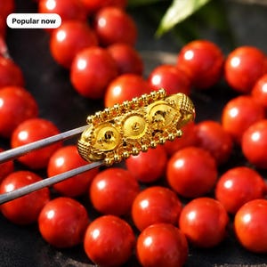 Può includere: Una perla cilindrica in oro con intricati dettagli in filigrana e granulazione, tenuta da una pinzetta metallica su uno sfondo di numerose perle rosse, lisce e rotonde. L'immagine include un'etichetta bianca con il testo Popular now.