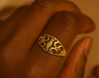 May include: A gold-colored ring with an oval face featuring an openwork design. The ring is worn on a finger, and the intricate pattern is visible against the skin. The ring's design is a series of swirling shapes.
