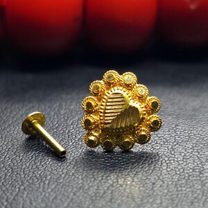 May include: A gold nose ring with a heart-shaped design. The heart is surrounded by a circle of small, round stones. The nose ring is attached to a small, gold post.