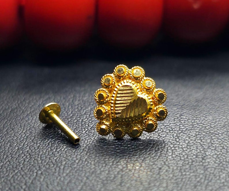 May include: A gold nose ring with a heart-shaped design. The heart is surrounded by a circle of small, round stones. The nose ring is attached to a small, gold post.