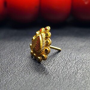 May include: A gold nose ring with a heart-shaped design and intricate details. The ring is set with a small red stone.