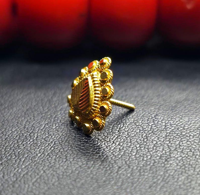May include: A gold nose ring with a heart-shaped design and intricate details. The ring is set with a small red stone.