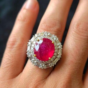 Puede incluir: Un anillo llamativo con un rubí ovalado de color rojo intenso, rodeado por un halo de diamantes baguette y redondos. El anillo tiene detalles dorados y una montura plateada. Una joya lujosa.