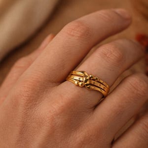Puede incluir: Un anillo dorado con un diseño único. El anillo presenta tres bandas con pequeños detalles redondos y un elemento central texturizado. El anillo es de color oro cálido y parece estar hecho de metal.