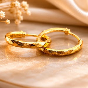 May include: A pair of gold hoop earrings featuring a textured, hammered surface finish. The circular earrings are linked together, showcasing a reflective metallic sheen and a distinct faceted pattern across the entire outer surface of the metal bands.