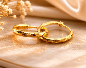 May include: A pair of gold hoop earrings featuring a textured, hammered surface finish. The circular earrings are linked together, showcasing a reflective metallic sheen and a distinct faceted pattern across the entire outer surface of the metal bands.