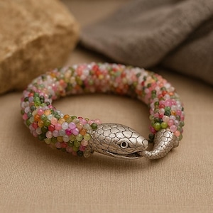 Könnte beinhalten: Ein Armband mit Schlangenmuster. Das Armband besteht aus kleinen, bunten Perlen in Rosa-, Grün-, Orange- und Gelbtönen. Der Kopf und der Schwanz der Schlange sind aus silberfarbenem Metall.