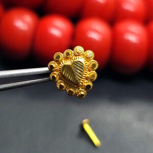 May include: A gold heart-shaped nose ring with a decorative border. The ring is held by a pair of tweezers.