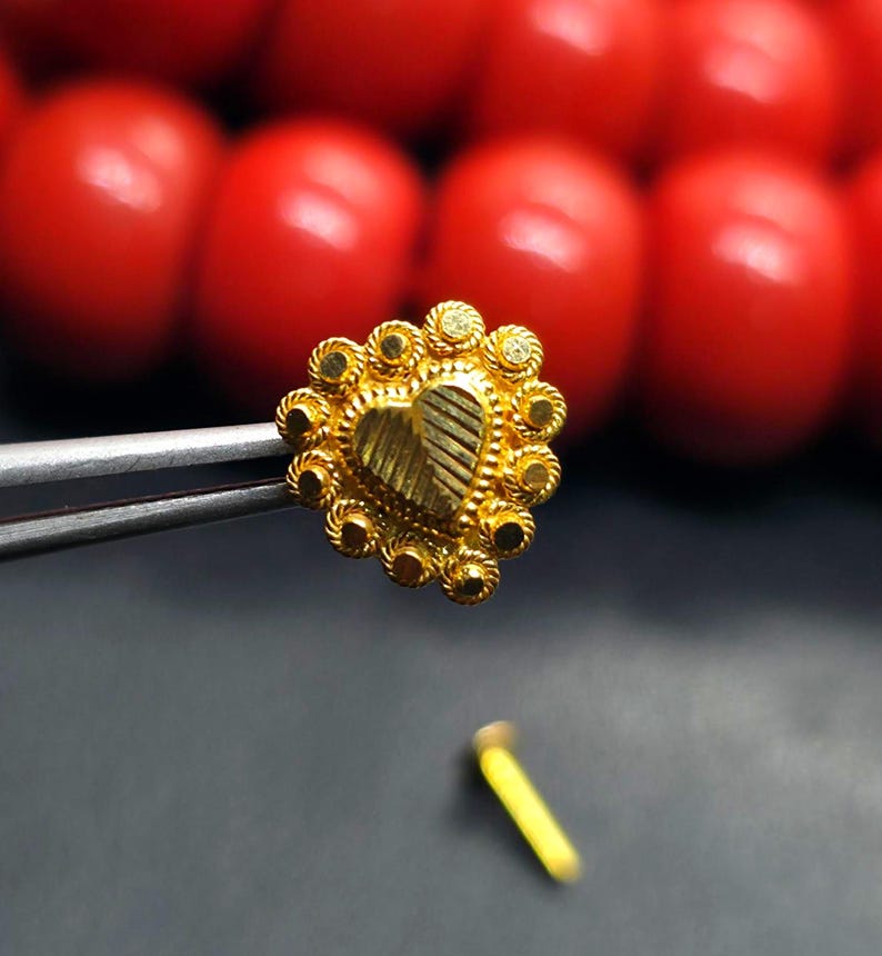 May include: A gold heart-shaped nose ring with a decorative border. The ring is held by a pair of tweezers.