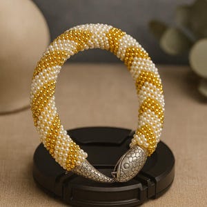 Pode incluir: Uma pulseira de contas com fileiras alternadas de contas brancas e douradas. A pulseira tem a forma de uma cobra, com um fecho de cabeça de cobra de metal prateado. A pulseira está sobre uma superfície preta.