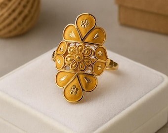 May include: A gold-colored ring with a floral design. The ring features a central flower surrounded by petal-shaped elements. The ring is displayed on a white surface, with a blurred background including a spool of twine and a box.