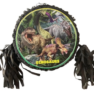 May include: A black and green dinosaur-themed pinata with black tassels. The pinata features a colorful image of four dinosaurs in a jungle setting. The text "DINOSAURS" is printed in yellow on the pinata.
