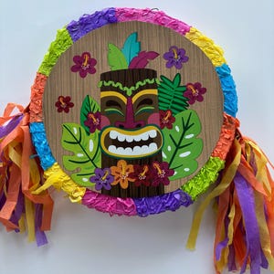 Könnte beinhalten: Eine farbenfrohe Piñata in Form eines Tiki-Kopfes mit einem lächelnden Gesicht. Die Piñata ist mit grünen Blättern, rosa Blumen und einem braunen Holz-Tiki-Kopf verziert. Die Piñata ist von bunten Seidenpapier-Streifen umgeben.