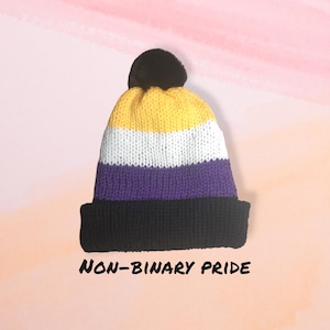 May include: A knitted beanie with a yellow, white, purple and black colour scheme. The beanie has a pom-pom on top and the text "NON-BINARY PRIDE" is written below it.