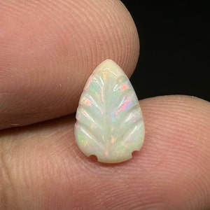 Könnte beinhalten: Ein geschnitzter Opal-Edelstein in Form eines Blattes. Der Opal hat eine cremeweiße Grundfarbe mit schillernden Blitzen in Rosa, Grün und Blau. Das Blattdesign weist detaillierte Adern und eine abgerundete Spitze auf. Der Edelstein wird zwischen Fingern gehalten.