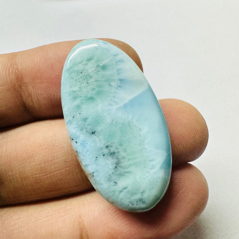 May include: A light blue oval cabochon gemstone with a cloudy, marbled pattern and dark gray inclusions.