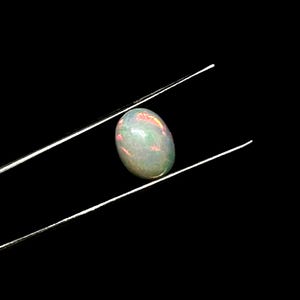 Puede incluir: Una piedra preciosa de &oacute;palo cabuj&oacute;n de forma ovalada sostenida por unas pinzas de metal plateado. La piedra presenta una base blanca lechosa transl&uacute;cida con destellos internos de luz iridiscente verde, naranja y rosa sobre un fondo negro.