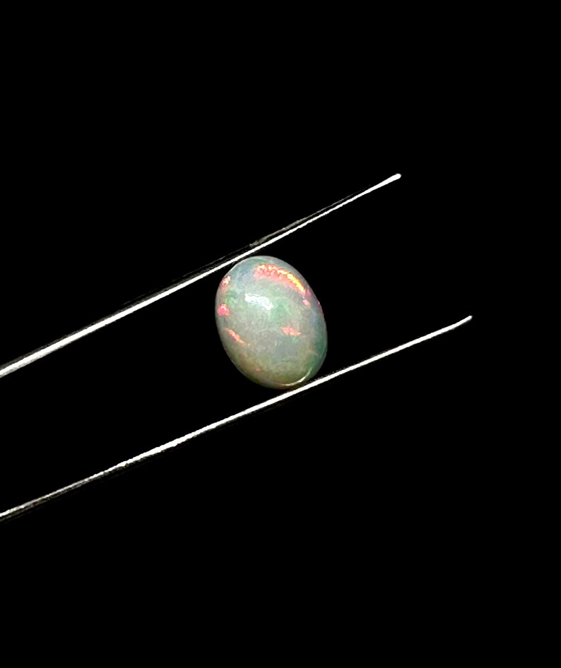 Puede incluir: Una piedra preciosa de &oacute;palo cabuj&oacute;n de forma ovalada sostenida por unas pinzas de metal plateado. La piedra presenta una base blanca lechosa transl&uacute;cida con destellos internos de luz iridiscente verde, naranja y rosa sobre un fondo negro.