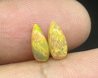 Puede incluir: Dos opales en forma de lágrima con un brillo iridiscente amarillo y verde. Las opales están colocadas sobre un fondo marrón claro.