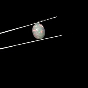 Puede incluir: Una piedra preciosa de &oacute;palo cabuj&oacute;n de forma ovalada sostenida por unas pinzas met&aacute;licas finas. La piedra presenta un color base blanco transl&uacute;cido con destellos internos de luz roja, verde y azul, creando un efecto de juego de colores.