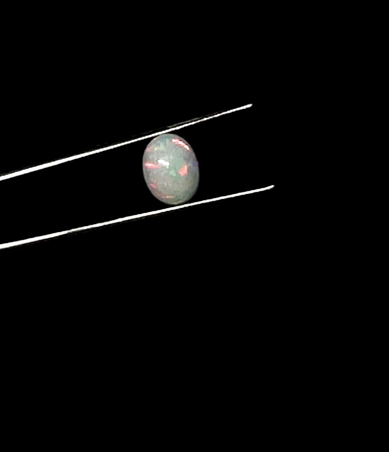 Puede incluir: Una piedra preciosa de &oacute;palo cabuj&oacute;n de forma ovalada sostenida por unas pinzas met&aacute;licas finas. La piedra presenta un color base blanco transl&uacute;cido con destellos internos de luz roja, verde y azul, creando un efecto de juego de colores.