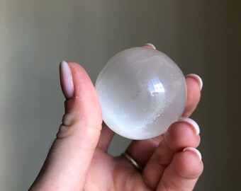 Op de afbeelding: Een witte selenietbol, een gladde, doorschijnende kristallen bol, vastgehouden in een hand.