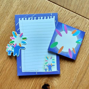 Puede incluir: Un bloc de notas azul con una página blanca rayada y un borde azul. El bloc de notas tiene una pegatina que dice "A heart is a heavy burden". Hay una pegatina colorida de un personaje de dibujos animados en el bloc de notas. El bloc de notas está sobre una superficie de madera.