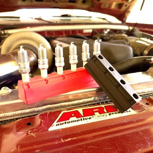 以下が含まれることがあります： 赤と黒のスパークプラグホルダーに5つのスパークプラグが入っています。ホルダーは車のエンジンの上に置かれています。車のエンジンには銀色のエアフィルターと黒いラジエーターがあります。