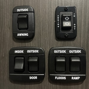May include: A panel of black switches with white labels. The switches control an awning, door, floods, and ramp. The labels read "Outside Awning", "Inside Outside Door", "Outside Floods", and "Outside Ramp".