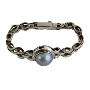 Peut inclure: Un bracelet en argent avec une perle grise irisée au centre. Le bracelet présente un motif en forme de chaîne avec des découpes ovales. Le fermoir est une barre rectangulaire simple. Le bijou est présenté sur fond blanc.