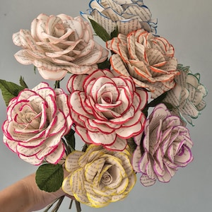 May include: A bouquet of seven handcrafted paper roses made from printed book pages. Each rose features distinctively colored petal edges in shades of pink, red, yellow, purple, orange, blue, and green, with dark green crepe paper leaves attached.