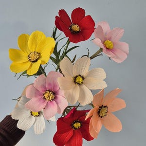 May include: A bouquet of eight paper flowers in various colors, including red, pink, yellow, and peach. The flowers are arranged in a hand, with the stems pointing upwards.