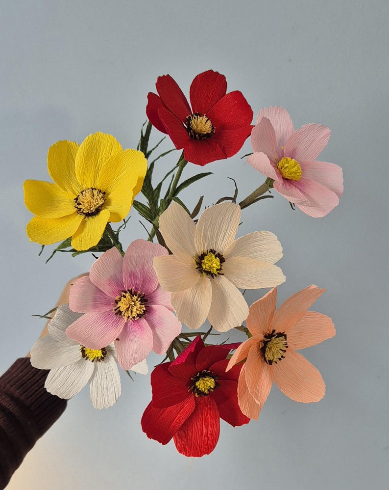 May include: A bouquet of eight paper flowers in various colors, including red, pink, yellow, and peach. The flowers are arranged in a hand, with the stems pointing upwards.