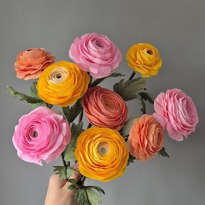May include: A vibrant bouquet of paper flowers in pink, orange, and yellow. The flowers feature a layered petal design and are held together by green paper stems and leaves. The background is a plain grey.
