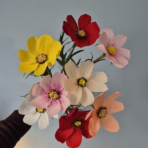 May include: A bouquet of paper flowers in various colors, including red, yellow, pink, and white. The flowers are arranged in a cluster and have a delicate, layered appearance.