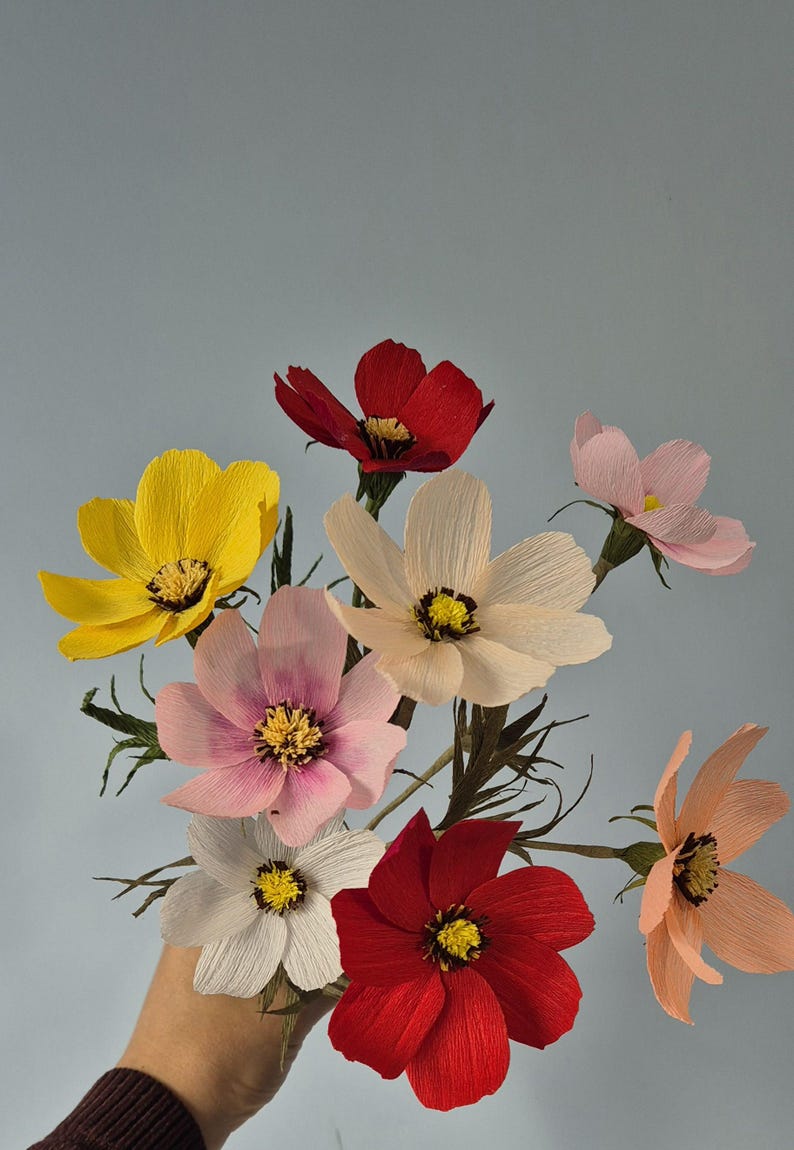 May include: A bouquet of paper flowers in various colors, including yellow, pink, red, and white. The flowers are arranged in a hand, with the stems extending out.