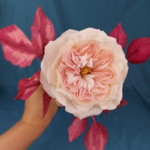 May include: A delicate pink and white paper rose with deep pink paper leaves. The rose is held in a hand against a blue background.