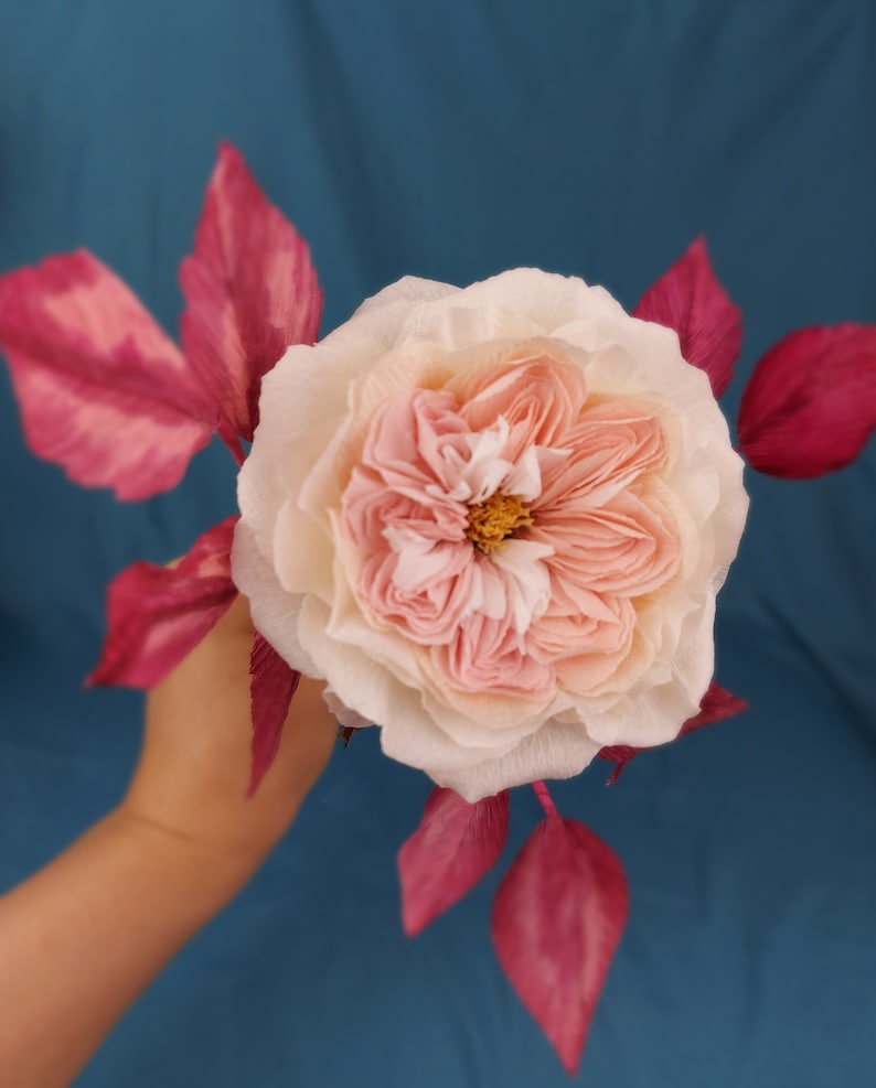 May include: A delicate pink and white paper rose with deep pink paper leaves. The rose is held in a hand against a blue background.