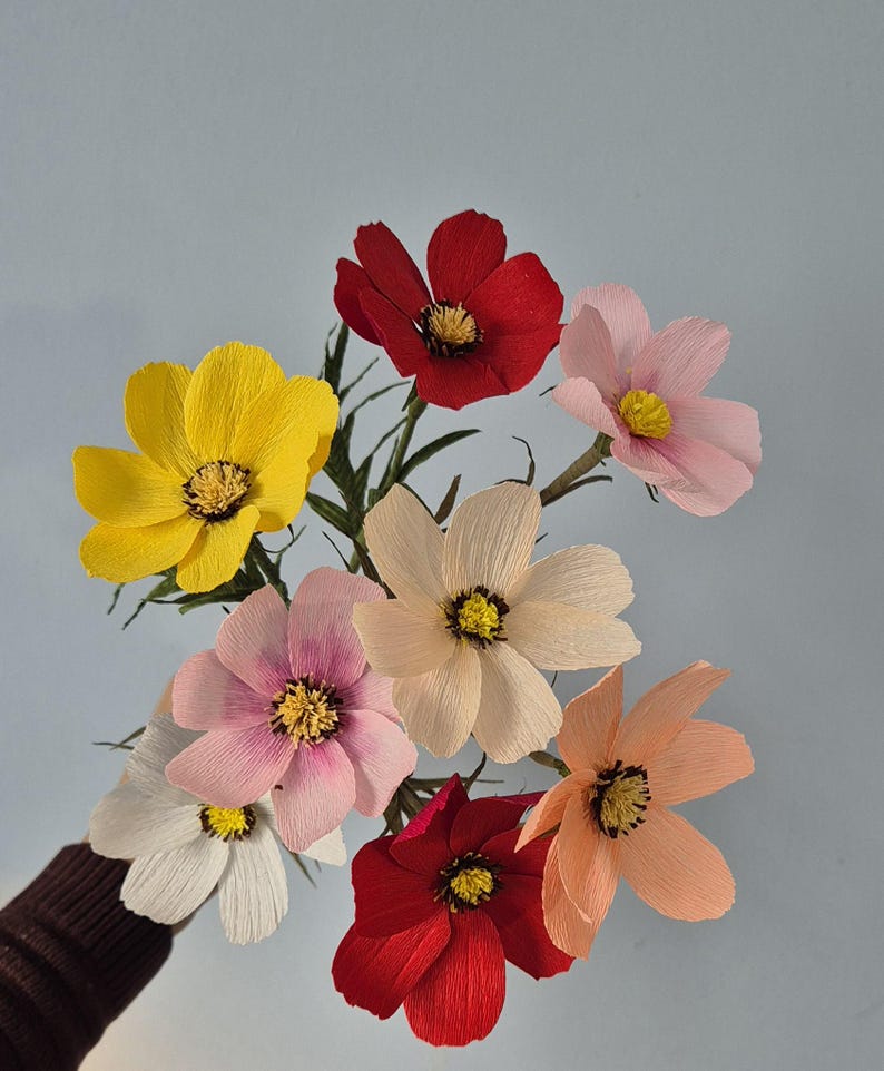 May include: A bouquet of seven paper flowers in various colors, including yellow, pink, red, and white. The flowers are arranged in a fan shape and have a delicate, layered appearance.