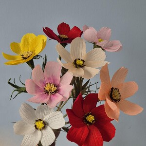 May include: A bouquet of seven paper flowers in various colors, including yellow, pink, red, and peach. The flowers are arranged in a hand, with the stems extending downwards.