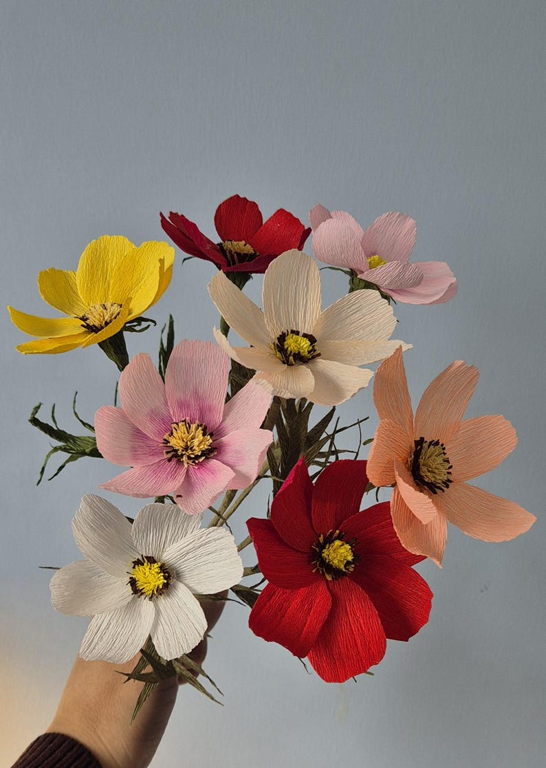 May include: A bouquet of seven paper flowers in various colors, including yellow, pink, red, and peach. The flowers are arranged in a hand, with the stems extending downwards.