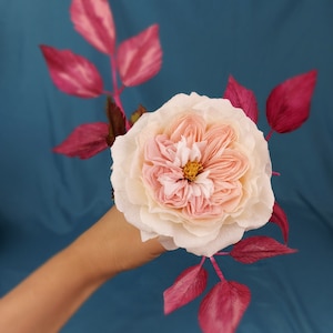 May include: A delicate paper rose with a blush pink and cream color scheme, accented by deep pink paper leaves. The rose is held in a hand against a teal blue background.