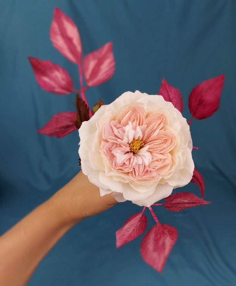 May include: A delicate paper rose with a blush pink and cream color scheme, accented by deep pink paper leaves. The rose is held in a hand against a teal blue background.