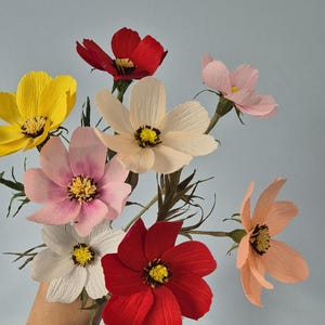 May include: A bouquet of seven paper flowers in various colors, including yellow, pink, red, and white. The flowers are arranged in a hand, with the stems extending out.